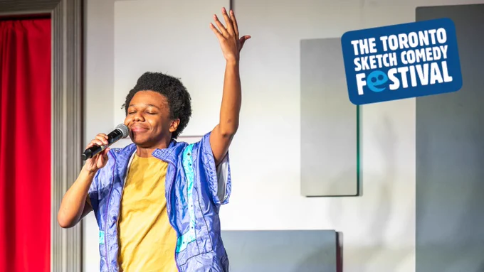Image Description: Ajahnis Charley stands onstage in a power pose with their eyes closed, a microphone in one hand, and their arm raised to the sky. The Toronto Sketch Comedy Festival logo appears in the top right corner