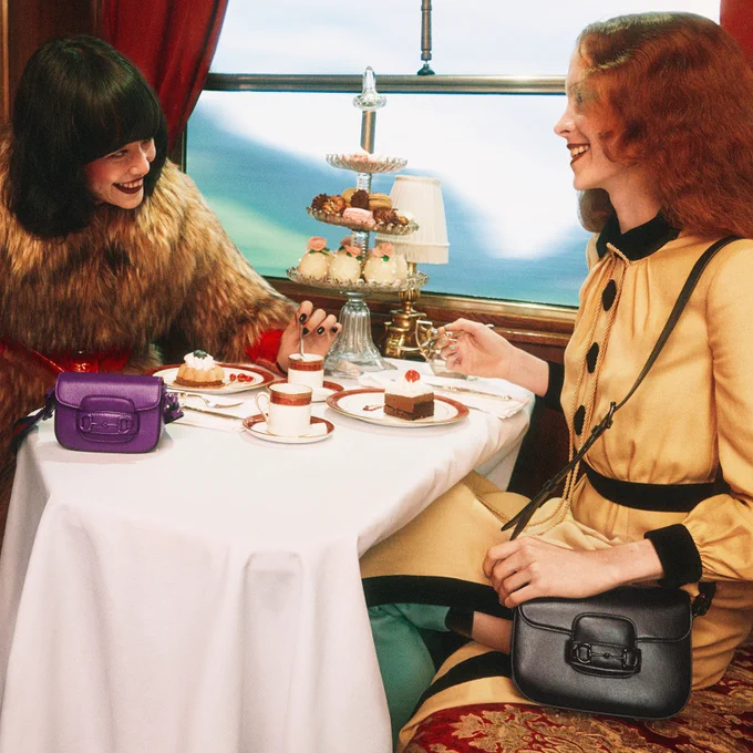 With a mountain landscape seen through the window of a vintage train car, an Asian woman in a faux fox fur coat smiles widely and stirs her espresso, meeting the smile of a white woman with long wavy red hair, seated across from her at a dining table set with several kinds of cake. On the table is a plum leather Horsebit 1955 mini bag with tonal enameled hardware and web strap, while the white woman, in a yellow silk dress, has a small black version of the bag over one shoulder. 