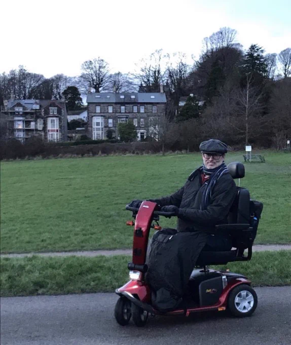Me on my mobility scooter with a waterproof fleecy lined BundleBean keeping my legs warm. Background is a park with stone faced Victorian house.