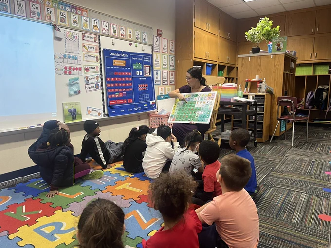 Teacher pointing to words on poster size AAC core board while children seated on carpet are watching.