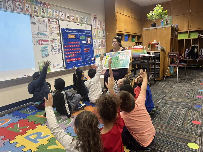 Teacher hold a poster size AAC core board with children seated on the floor with hands raised.