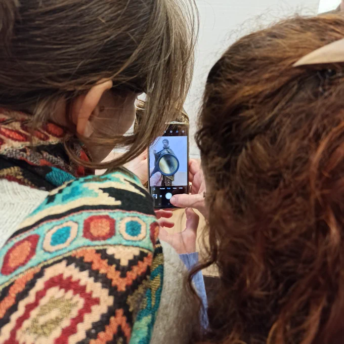 View over the shoulder of two students with dark hair examining a sculpture in front of them with a magnifying glass. One student is taking a picture with her smart phone.