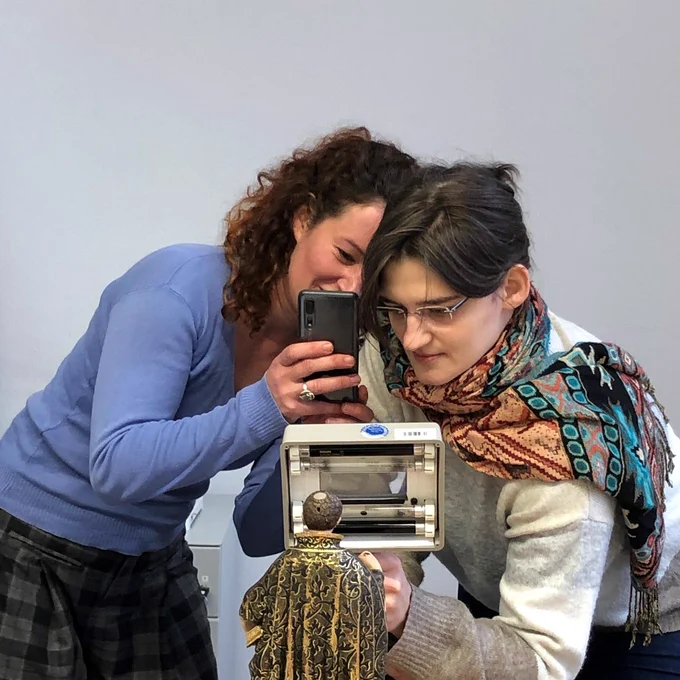 Two students are looking at a small sculpture in front of them. One ist holding a magnifying glass and the other is taking a picture of the scene with her smart phone.
