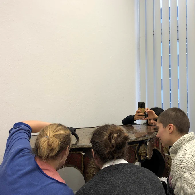 Four students seen from the back are kneeling around a furniture, closely examining its surface. One is taking a picture with her smartphone.