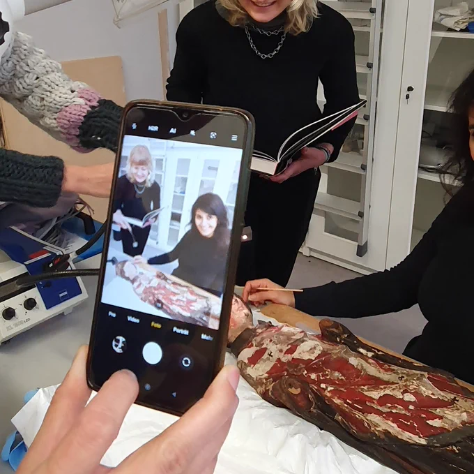 A smart phone is placed in the middle of the picture, capturing the scene behind. Two students are examining a sculpture placed on a table.