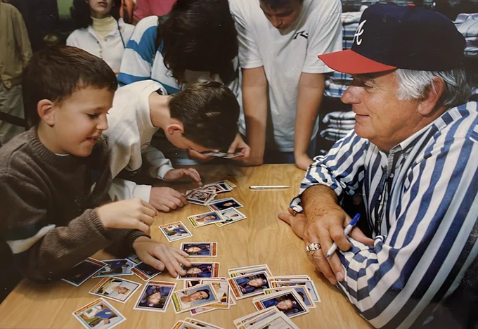 Gaylord Perry sitting at table surrounded by children of various ages looking at baseball cards laid out on the table.