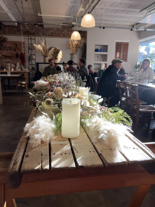 In the foreground, a candle stands on a rustic wooden table behind, it is a collection of autumn leaves, flowers, cones, attractively, decorating the table. In the background people sit together, enjoying food and drink the winter, sun shining gently through.