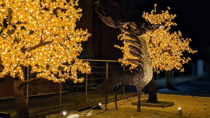 A deer like creature, woven from branches, stands proudly between the soft, warm, glow of two trees who's leaves are made from small fairy lights.