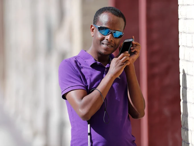 A man holds a smartphone up to his ear to listen to the voiceover. He holds a white cane under his other arm