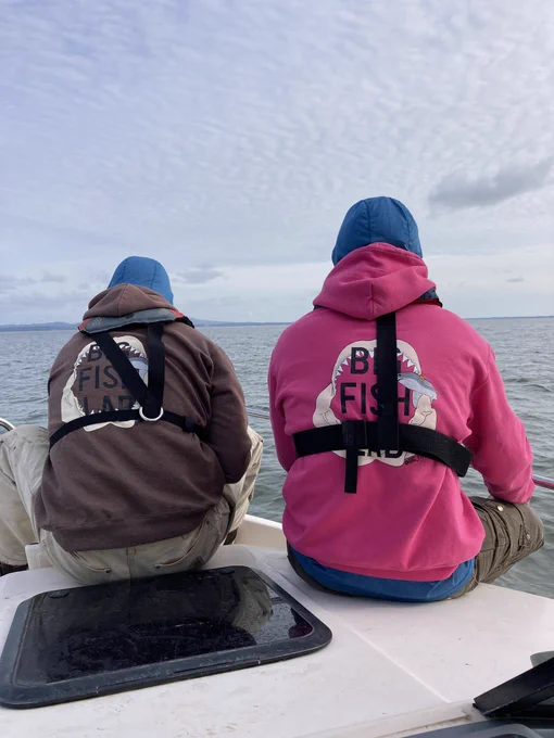Two Big Fish Lab members sitting at the front of a boat wearing big fish lab sweaters. One is brown and the other is hot pink!
