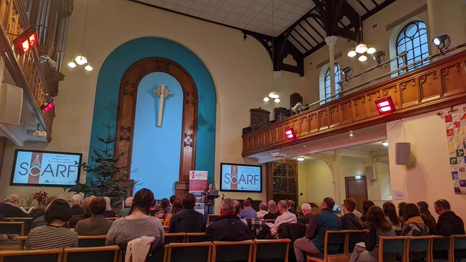 Conference guests listen to a speaker on a raised stage on an altar. 