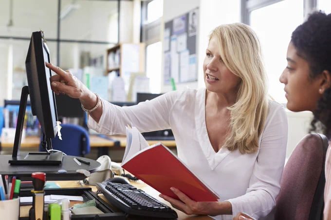 A student standing next to an educator, who is holding an open book while pointing to a computer screen.