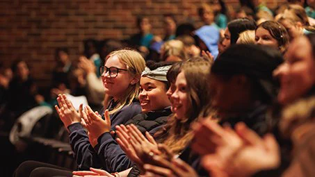 A row of young people in the audience smile and applaud. More young people can be seen behind them out of focus.