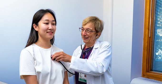 Clinician and patient in an exam room. Smiling patient sits on a treatment table while the smiling clinician listens to her heart with a stethoscope