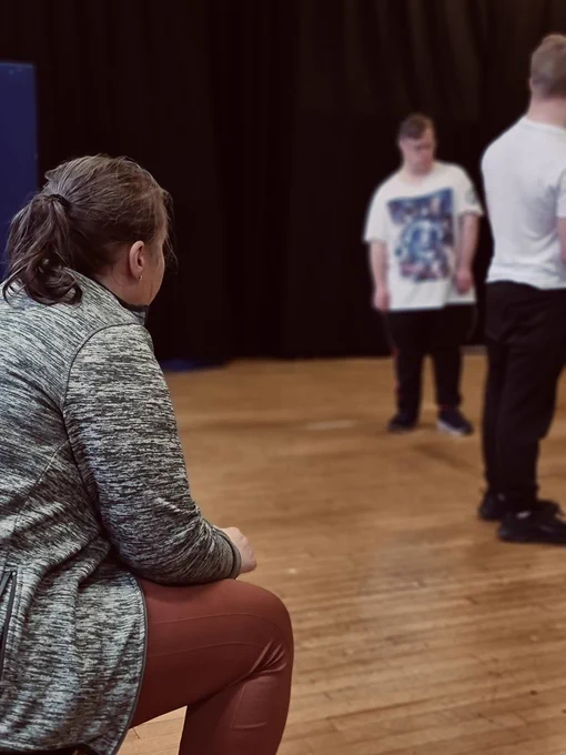 Foreground: The back and side of a seated person is in view - they are leaning forward engaged in the rehearsal in front of them. 
Out of focus background: Two performers interact with one another - it is unclear of their relationship. 