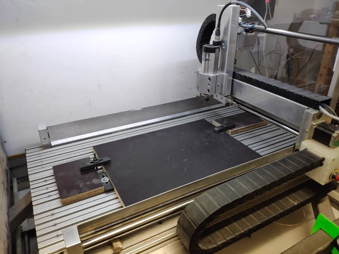 The picture shows a small flatbed CNC machine made from Aluminium profiles. In the working area a piece of dark plywood is mounted.