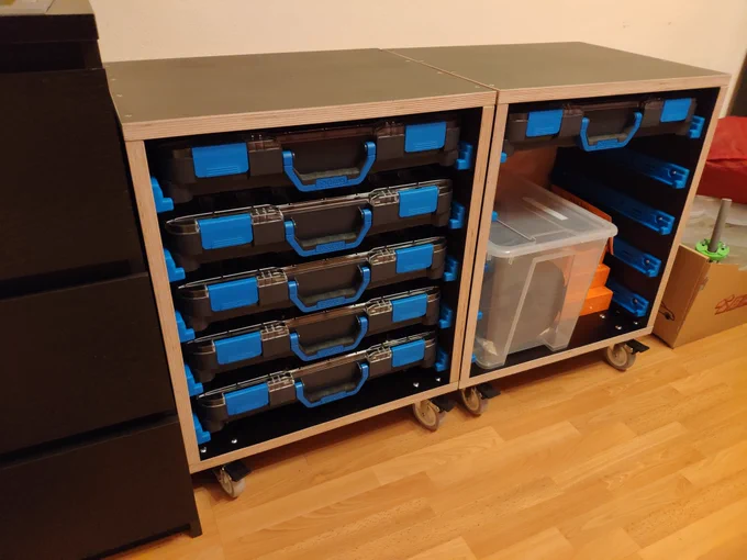 Two roughly square shapes racks made from thick dark plywood on castor wheels are standing in front of a wall. In one of them there are five identical boxes. The other one houses a sixth box at the top, revealing blue plastic rails on the side walls. A transparent plastic container is also in this rack.
