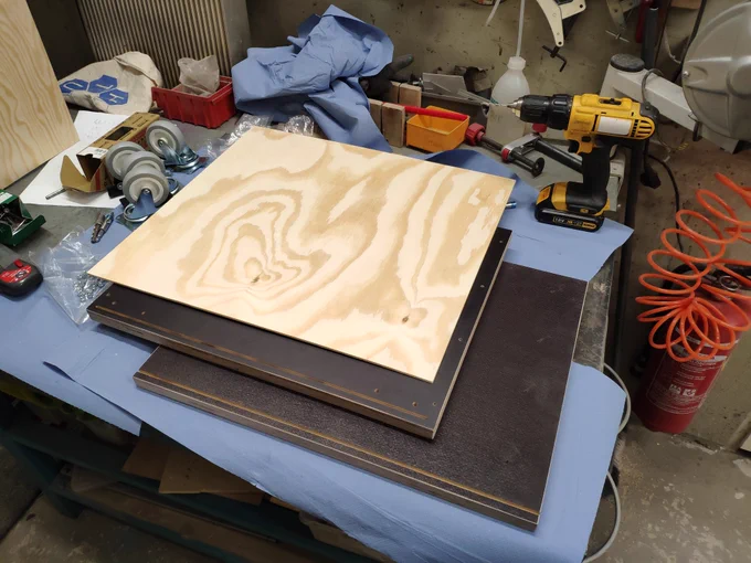 Multiple sheets of plywood are placed on top of each other on a workbench. One of them is thin and nicely textured. The oder ones are thick and dark. Also, they have holes and slots machined into them. Beside the plywood a cordless drill and a lot of tools is lying around.