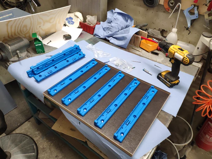 A dark, thick piece of plywood is lying on a workbench with some paper underneath it. Five blue plastic rails are mounted on top of it. More rails are lying next to it. Also there is a cordless drill. Screws and a lot of other stuff is lying on the workbench too.