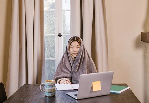 Someone sits at a brown wood table with a tan fleecy blanket wrapped around them and covering their hair. There is a mug, notebooks, and a silver laptop with a sticky note on it sitting on the table.