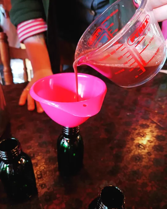 PHOTO of someone pouring red coloured herbal syrup through a bright pink funnel into a small brown glass bottle, on a table with other wee brown bottles