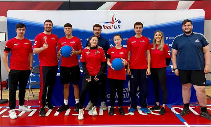 A group of volunteers stood posing for a photo wearing red Goalball UK tops. They are smiling for the camera and two of them are holding up goalballs too.
