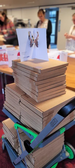 A stack of boxes on a trolley with a pop up card on top. People are working on the boxes in the background. 