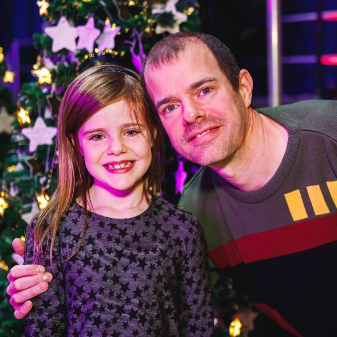 Man and young girl smiling at the camera in front of a decorated Christmas tree. 