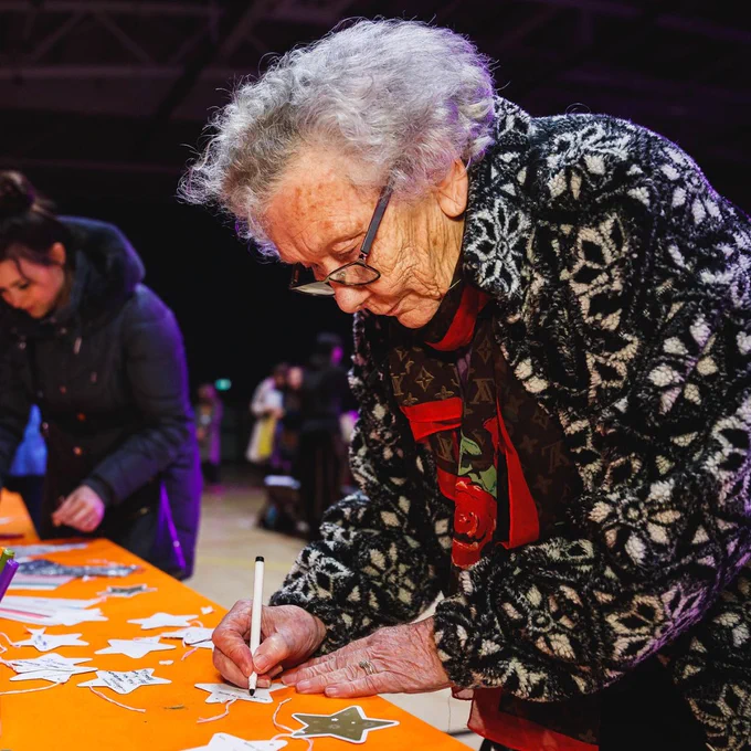 Elderly woman wearing a Christmas jacket and glasses, writing on a star decoration.