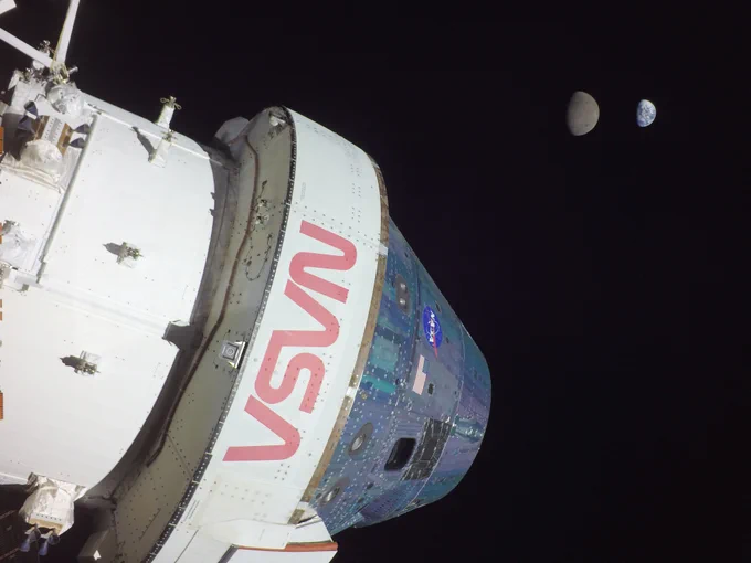 The Orion spacecraft stands out against the blackness of space. It shines bright with white, silver, and blue colors with a large red NASA logo in the middle. The Moon and Earth can be seen in the distance; the Moon is grey, and Earth is white and blue swirls. Credit: NASA