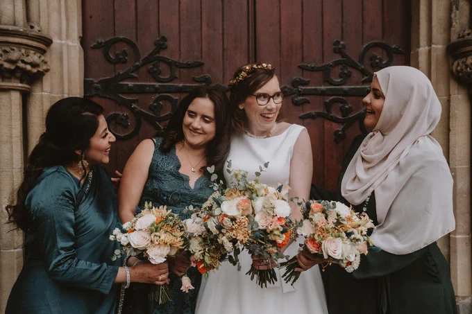 Four women are laughing against a backdrop of an ornate wooden door. One is wearing a simple sleeveless wedding dress and has amber flowers in her hair. The other three are in varying shades of teal. They are all carrying orange and cream bouquets.