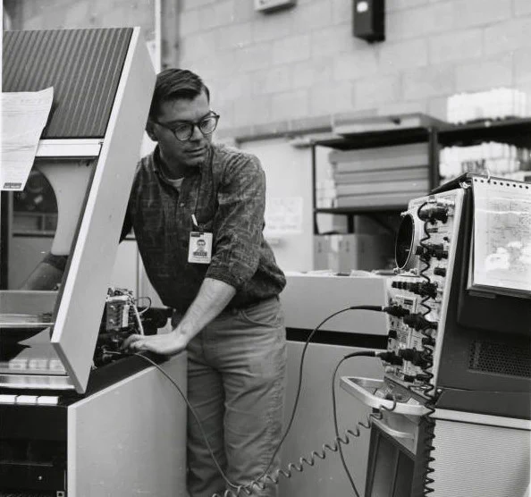 A maintenance worker tests mainframe equipment using digital testing equipment wired to the mainframe he is working on. Black and white square photo.