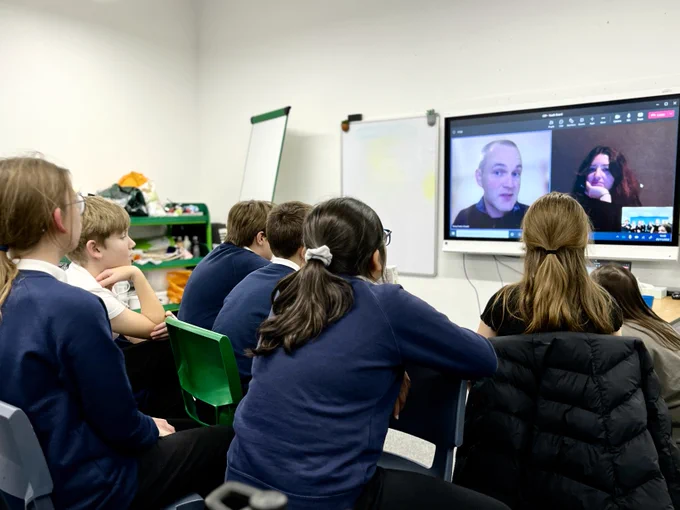 Children sit and speak to two people who are on a screen, connected via Zoom