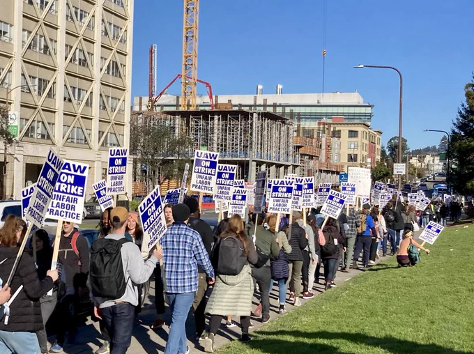 Workers holding picket signs reading "UAW On Strike Unfair Labor Practice" march along a sidewalk with large buildings and construction across the street and a green lawn in the foreground