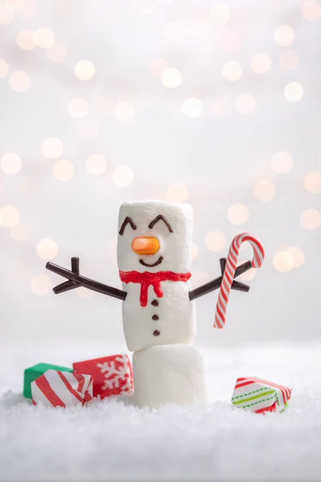 A snowman crafted from marsh mallows, with sweets as presents, on a bed of ice with festive lights in the background.