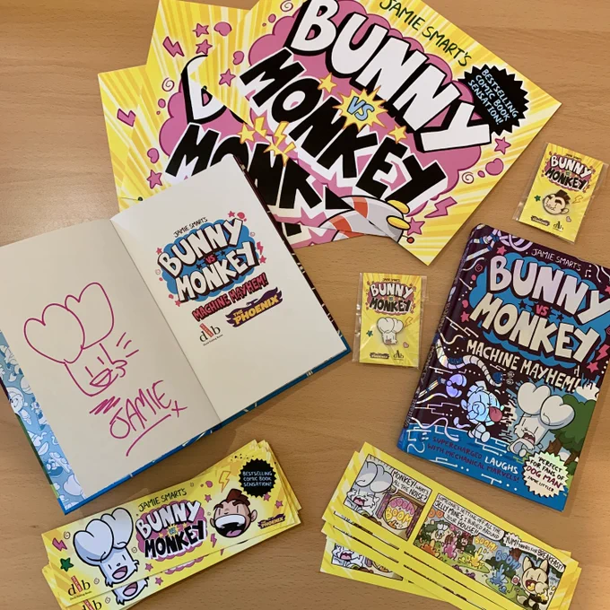 Bright yellow Bunny vs Monkey bookmarks and posters, two copies of Bunny vs Monkey: Machine Mayhem and some Bunny & Monkey pin badges are on a brown desk.