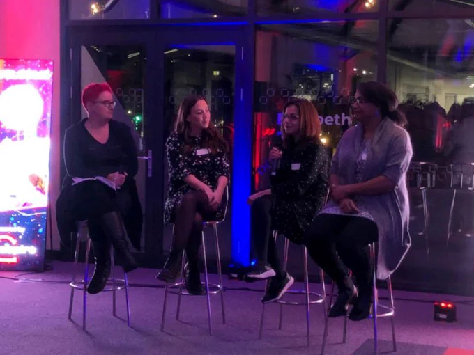 Image shows four women sitting on bar stools talking with microphones to an unseen audience.