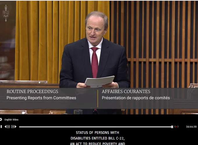 Picture of Chair of the HUMA Standing Committee in House of Commons, standing holding a text and speaking to the tabling of the Standing Committee's Report on C-22 with amendments.  On the screen is text entitled "Routine Proceedings - Presenting Reports from Committees" 