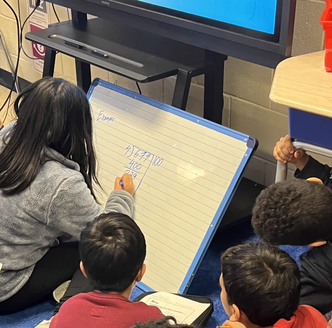 Student writing on white board with peers around her working together on one division problem using partial quotient.