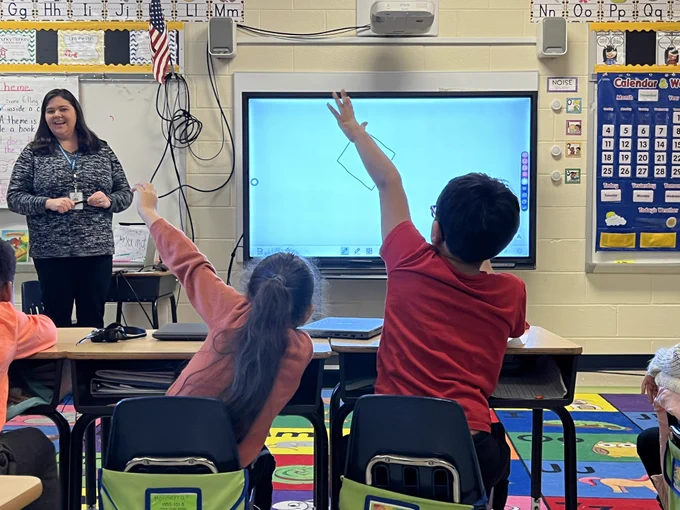 Teacher standing in front of room with students raising their hand.
