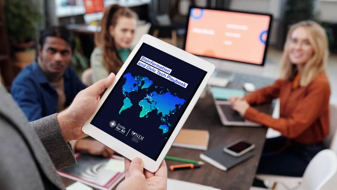 A person holds a tablet in front of a group of three people. The tablet screen reads: Disinformation and Civic Tech Playbook, with a world map and the Code for All and NDI logos.