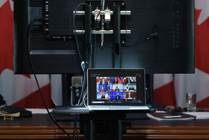A computer tablet is sitting on a shelf, which is in front of the desk in the Prime Minister’s West Block office. On the screen of the tablet, Prime Minister Justin Trudeau and other G7 leaders are seen in a virtual meeting. Behind the tablet, the back of a large monitor and a ring light are visible. In the background, four Canadian flags are visible.