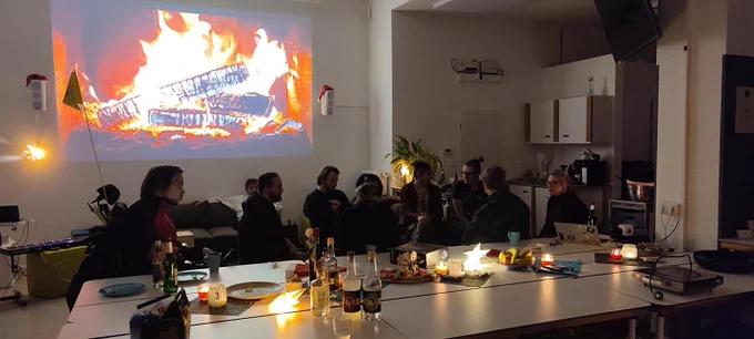 Das Team sitzt gemeinsam in einer Runde. Im Hintergrund ist auf der Wand ein virtuelles Kaminfeuer zu sehen.