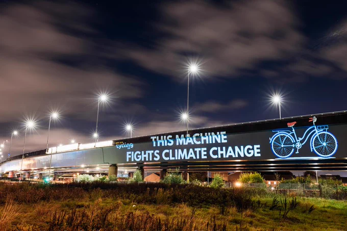 Projection on the side of a road bridge reads 'This machine fights climate change'. An image of a bike and the Cycling UK logo are also included.