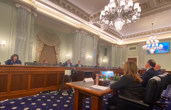 Room with wooden desk and four people behind it and 4 people in the foreground who are the witnesses