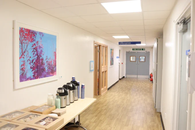 The Emmeline Centre corridor with a sign directing towards reception, a table of food and drinks and a framed piece of art on the wall with blue, pink and purple colours.