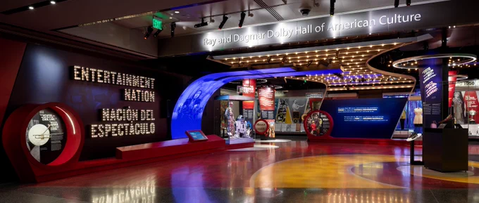 Entrance to "Entertainment Nation" exhibition, with title on wall in lights in English and Spanish. There's a spiral yellow path (like the yellow brick road) and Ruby Silppers, trumpet, and droids from Star Wars are slightly visible. Gallery includes text above: Ray and Dagmar Dolby Hall of American Culture.