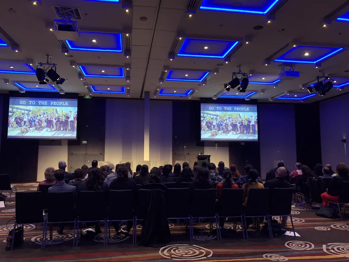 Conference room, view form the back of the room with people sitting in rows of chairs facing the front. At the front there are two screens showing a PowerPoint presentation. The slide on the screen says ‘Go To The People’, those words sit above a photo of a large group of people gathered for a group photo.