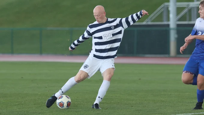 Matt McDonald wears a striped jersey and white shorts as he kicks a soccer ball on the field, his arms outstretched. 