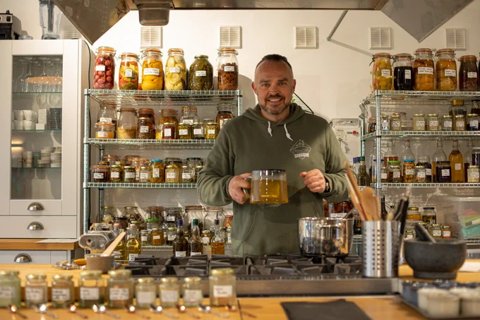 Ground up cookery. Man in the cookery school holding a jug of oil. Behind him on metal shelves are various cooking ingredients in labelled jars. in front is a stove with potted cooking utensils and more jarred ingredients. 